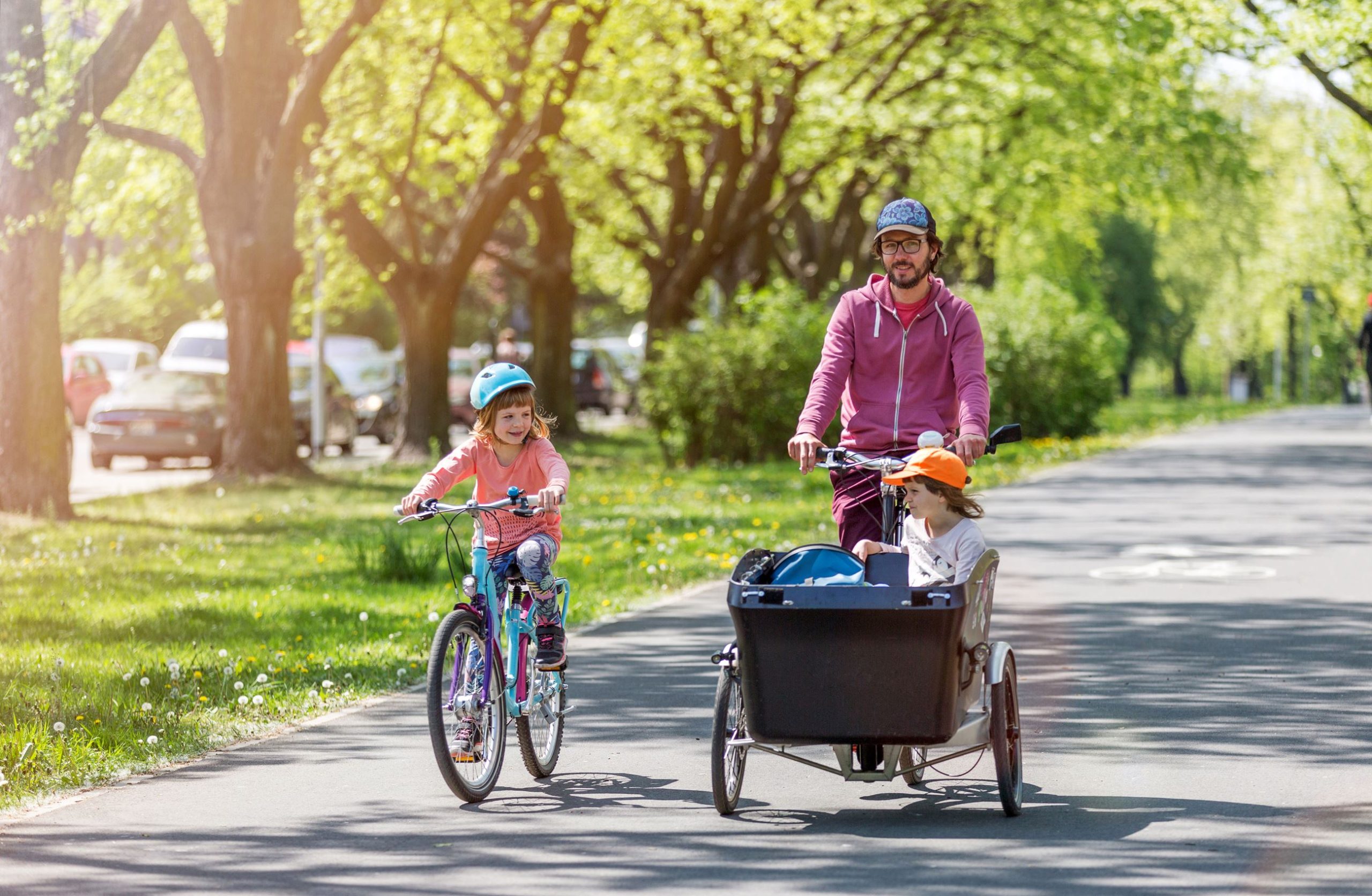 A father bicycling with two children — one riding alongside on her own bike, one riding in a cargo trailer — along a shaded, tree-lined path